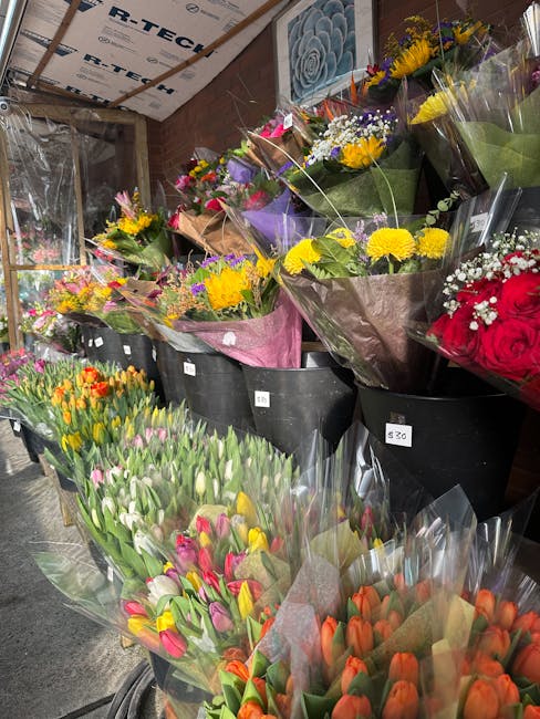 Indoor display of numerous flower bouquets in a florist shop, arranged on black plastic containers and a table. The bouquets feature vibrant, fresh flowers such as yellow daisies, purple and white tul