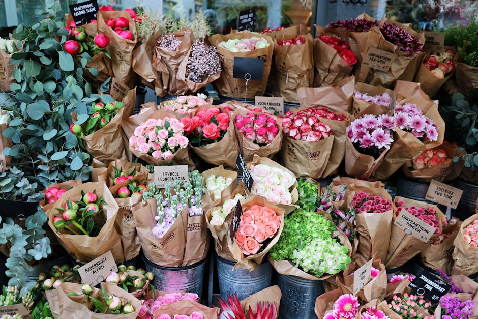 A vibrant display of freshly cut flowers arranged in brown paper wrapping, including pink roses, white and pink daisies, purple lisianthus, and green foliage. The bouquets are placed on the floor and